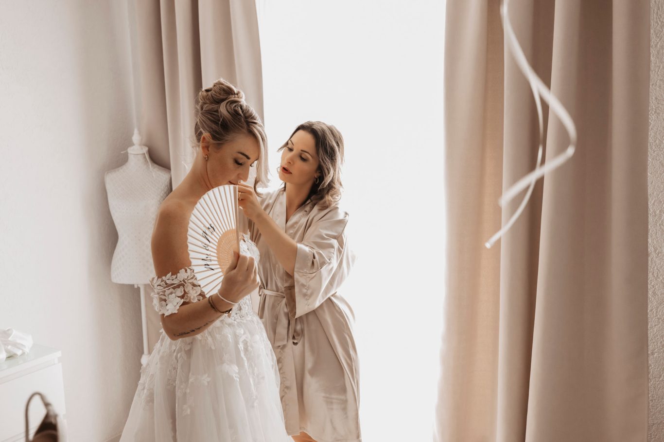 Braut wird von einer Frau beim Anziehen des Kleides in einem hellen Raum unterstützt.