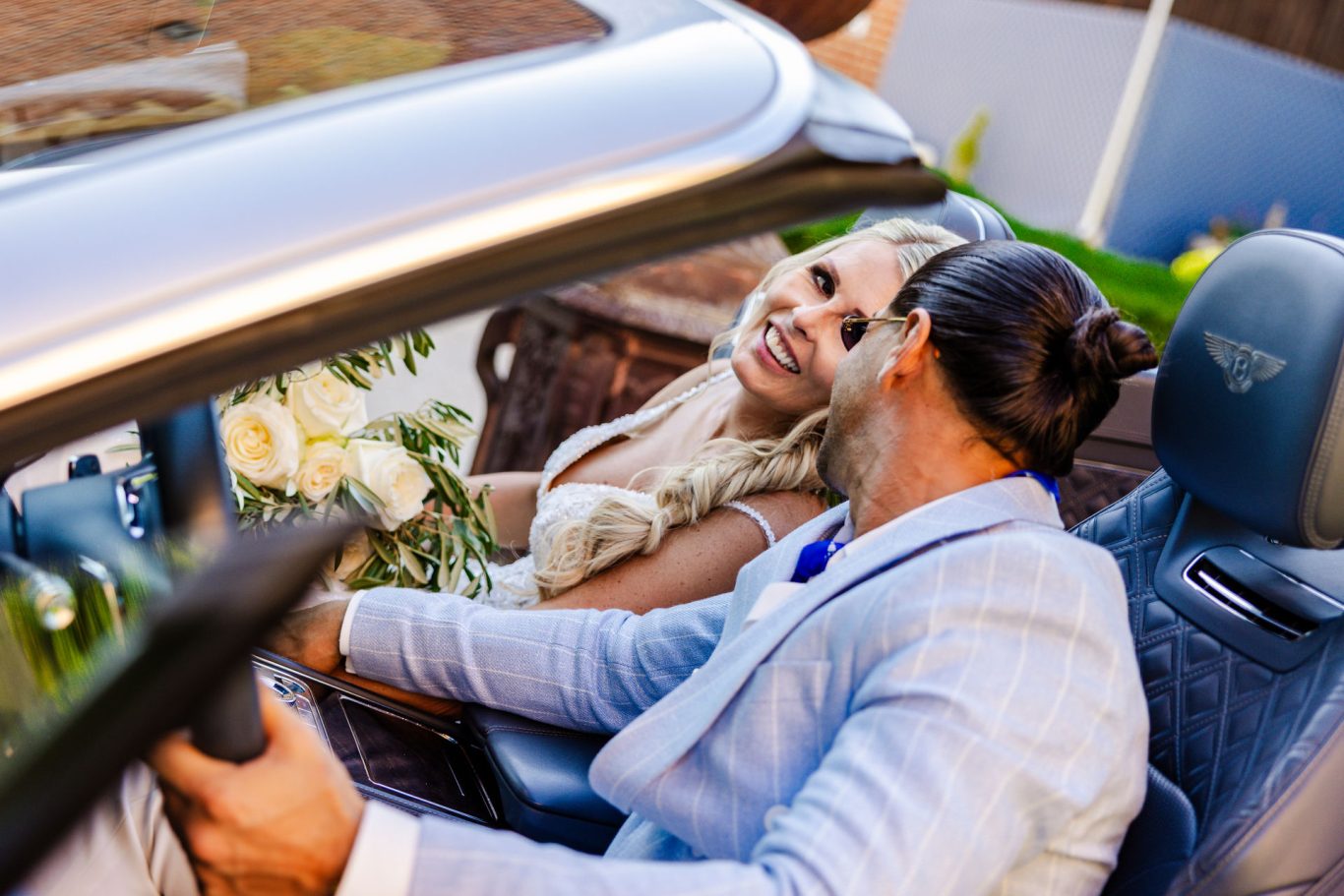 Braut und Bräutigam sitzen lachend in einem Cabrio mit Blumenstrauß.