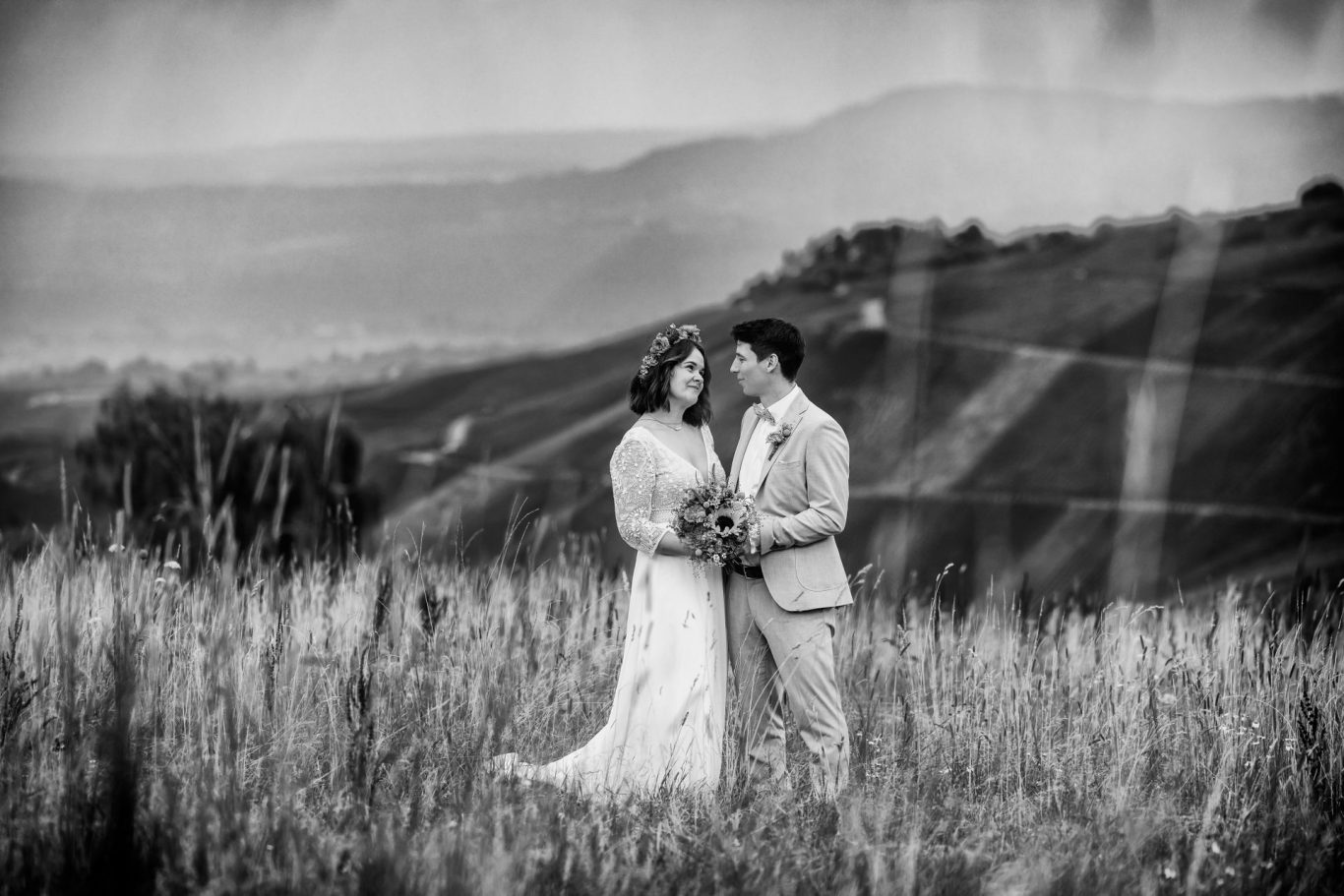 Ein Paar in Hochzeitskleidung umarmt sich in einer malerischen, bewölkten Landschaft.