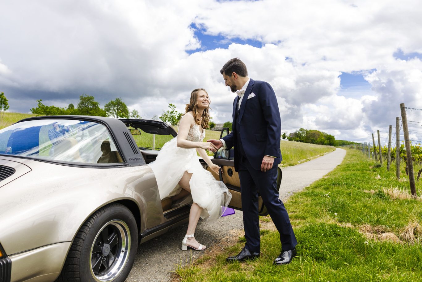 Hochzeitspaar an einem Porsche auf einem Weg zwischen Weinreben.