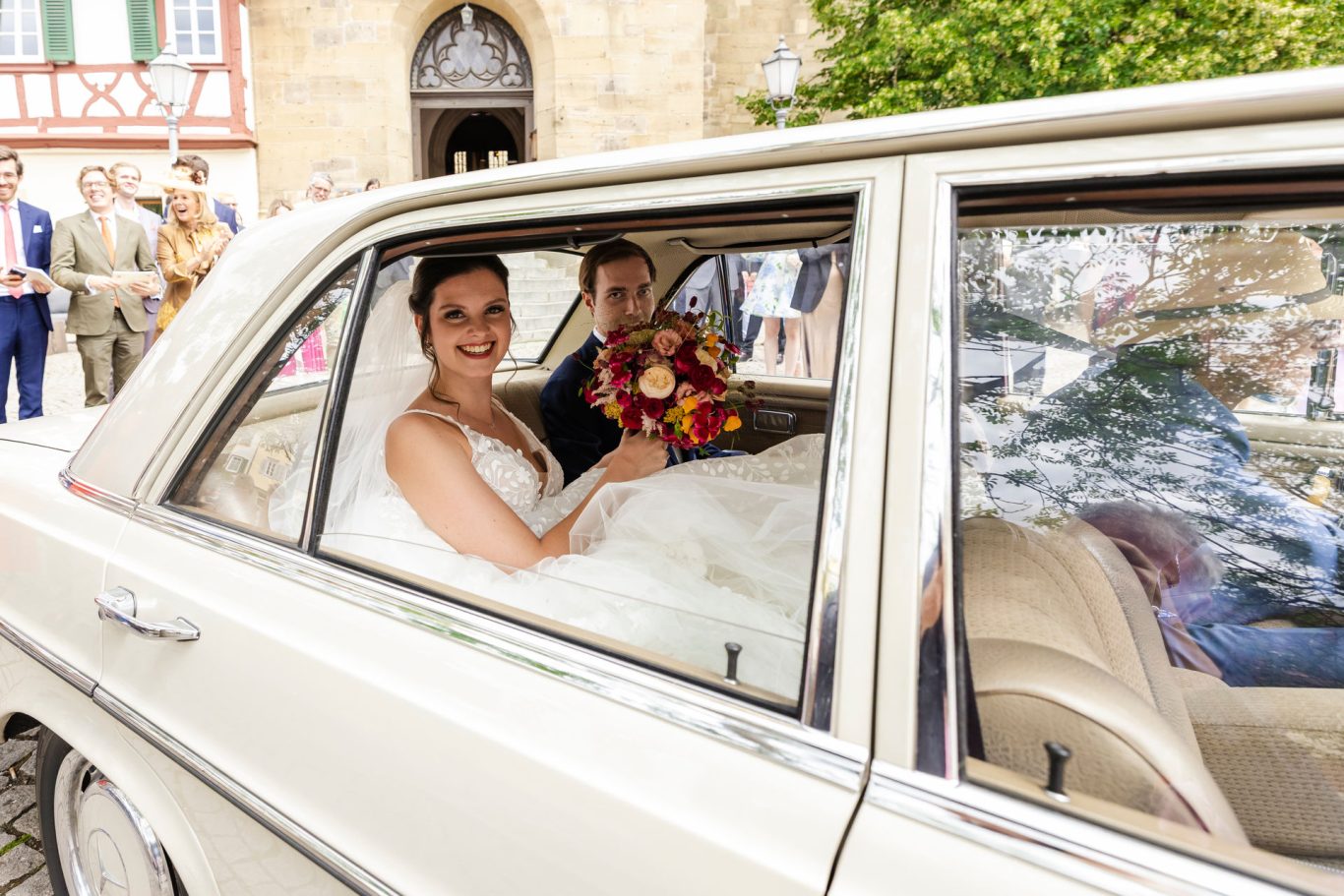 Hochzeitsfotograf Öhringen. Braut und Bräutigam sitzen lächelnd in einem Auto, umgeben von Hochzeitsgästen.