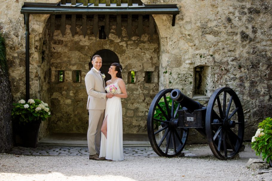 Paar in Hochzeitskleidung vor einer historischen Mauer mit einer Kanone.