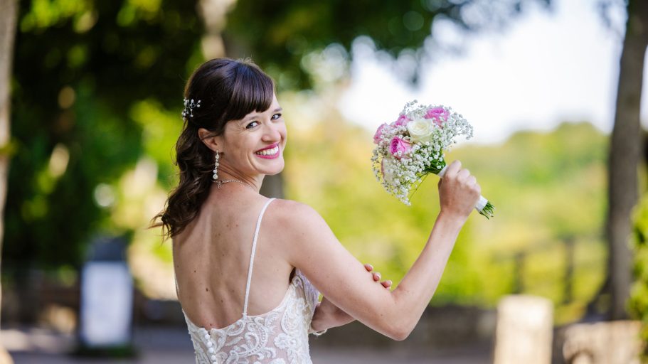 Braut mit Blumenstrauß, lächelnd und mit freiem Rücken, im Freien vor grüner Kulisse.