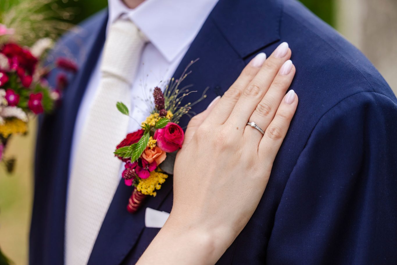 Ein Hand mit Ring berührt den Anzugkragen, umgeben von bunten Blütenarrangements.