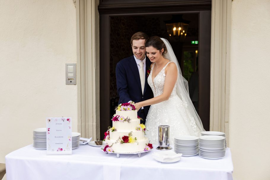 Schlosshotel Friedrichsruhe - Braut und Bräutigam schneiden eine Hochzeitstorte mit BlumenDekoration.