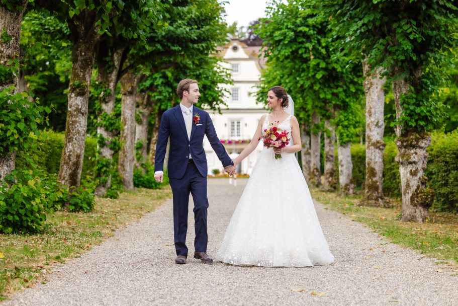 Vor den Schloss Friedrichsruhe - Brautpaar geht Hand in Hand auf einem Weg, umgeben von Bäumen.