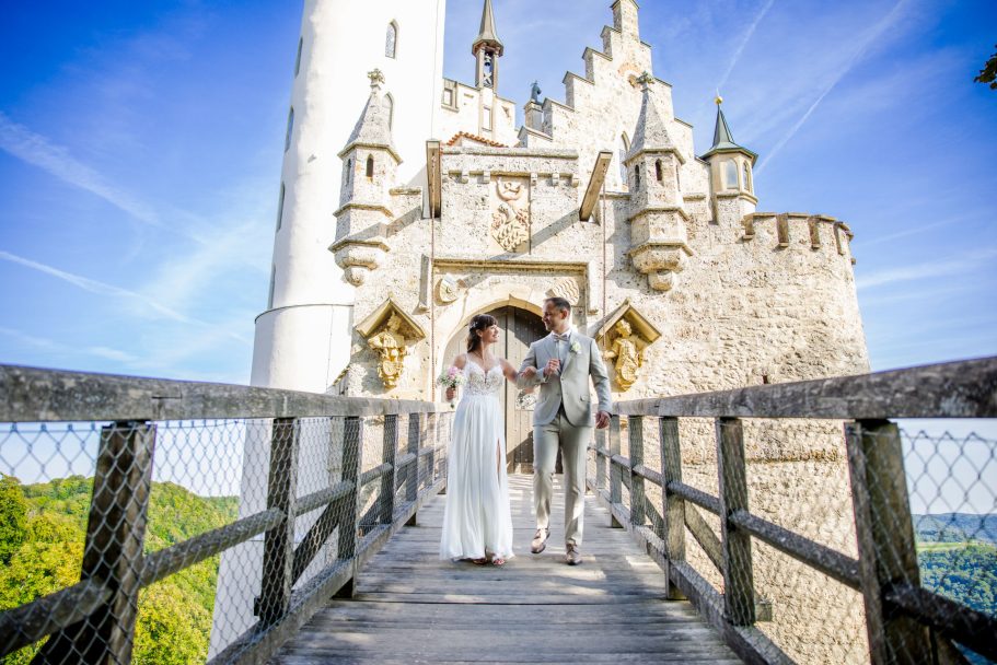 Paar in Hochzeitkleidung über eine Holzbrücke vor einem Schloss mit Türmen.