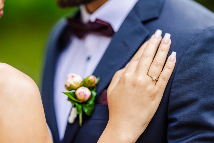 Hochzeitsdetails: Hand mit Ring, Anstecker am Anzug und eleganter Kleidung.